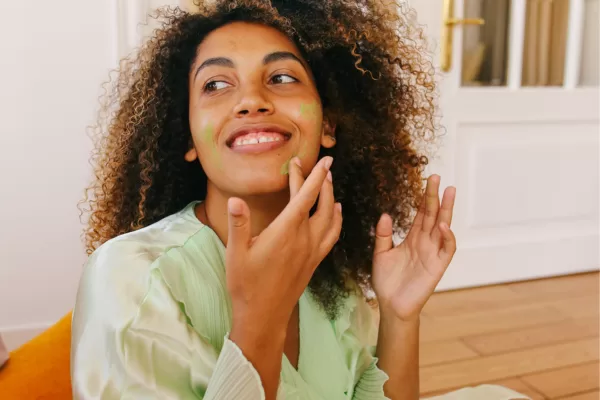 Mulher aplicando máscara facial natural verde, sorrindo e curtindo momento de autocuidado em ambiente iluminado.