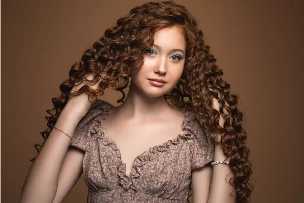 Mulher jovem com cabelo ruivo cacheado longo, maquiagem suave e vestido floral em fundo marrom quente.