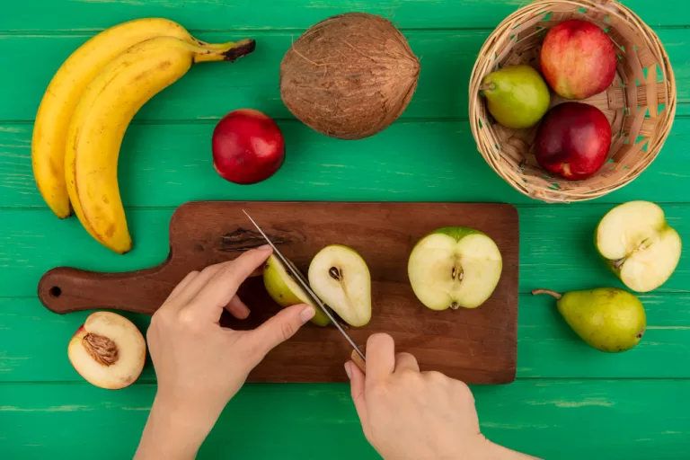 Mãos cortando maçã verde em tábua com frutas naturais ao redor, como banana, pêssego, coco e pera.
