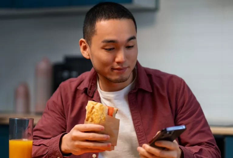 Homem comendo sanduíche enquanto usa celular à mesa com suco de laranja em ambiente doméstico.